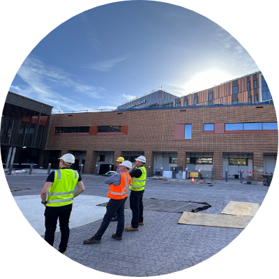 Three men wearing safety vests stand in front of a building, engaged in discussion about the construction project